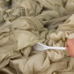 A hand uses a white spoon to stir tan sheets soaking in cloudy laundry stripping water.