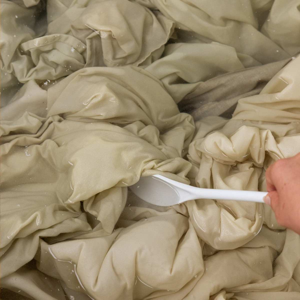 A hand uses a white spoon to stir tan sheets soaking in cloudy laundry stripping water.