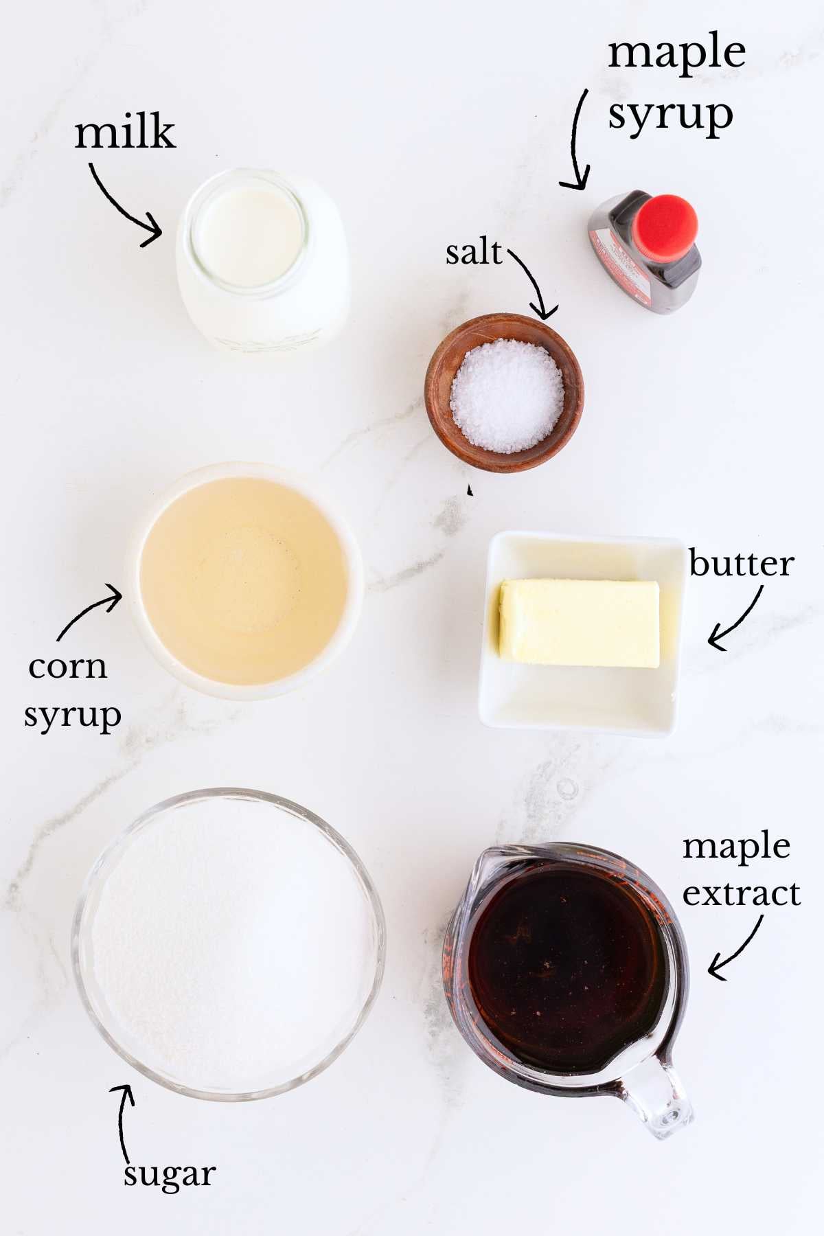 Labeled ingredients for maple caramels including milk, sugar, corn syrup, butter, salt, maple syrup, and maple extract on marble.