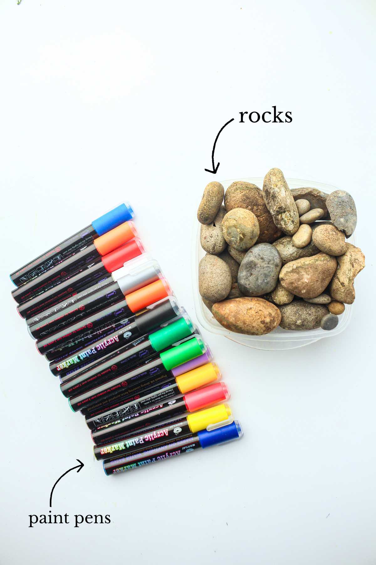 A row of colorful acrylic paint markers beside a plastic container filled with smooth natural rocks.