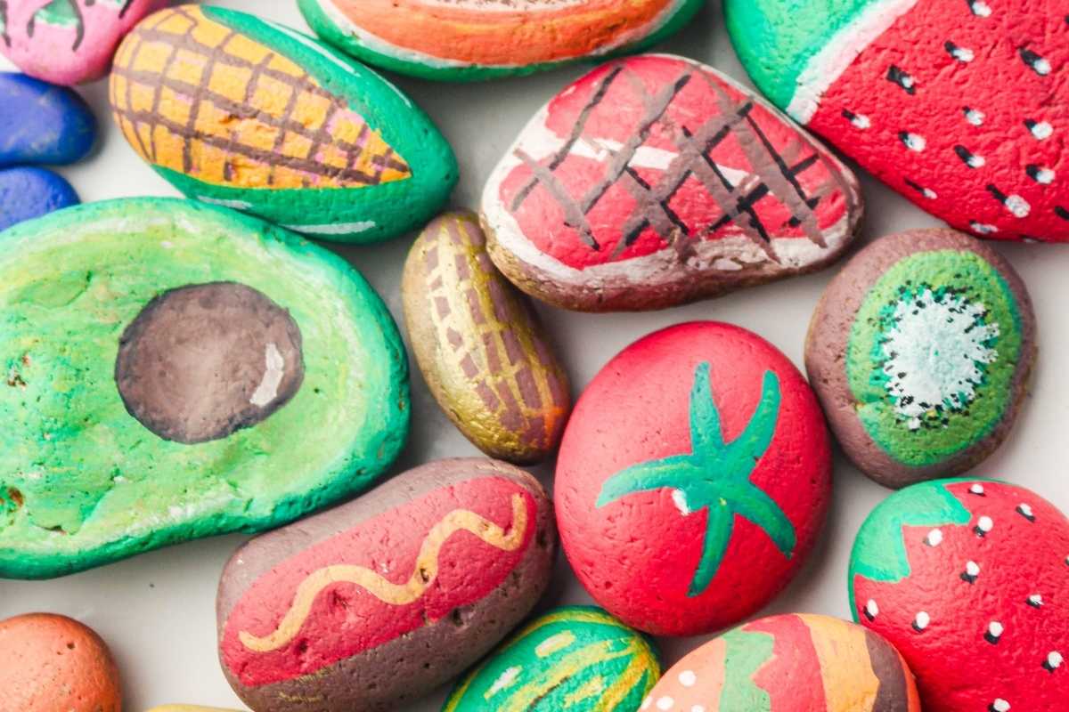 Colorful painted rocks resembling avocado, corn, watermelon, tomato, strawberry, kiwi, and hot dog arranged closely together.