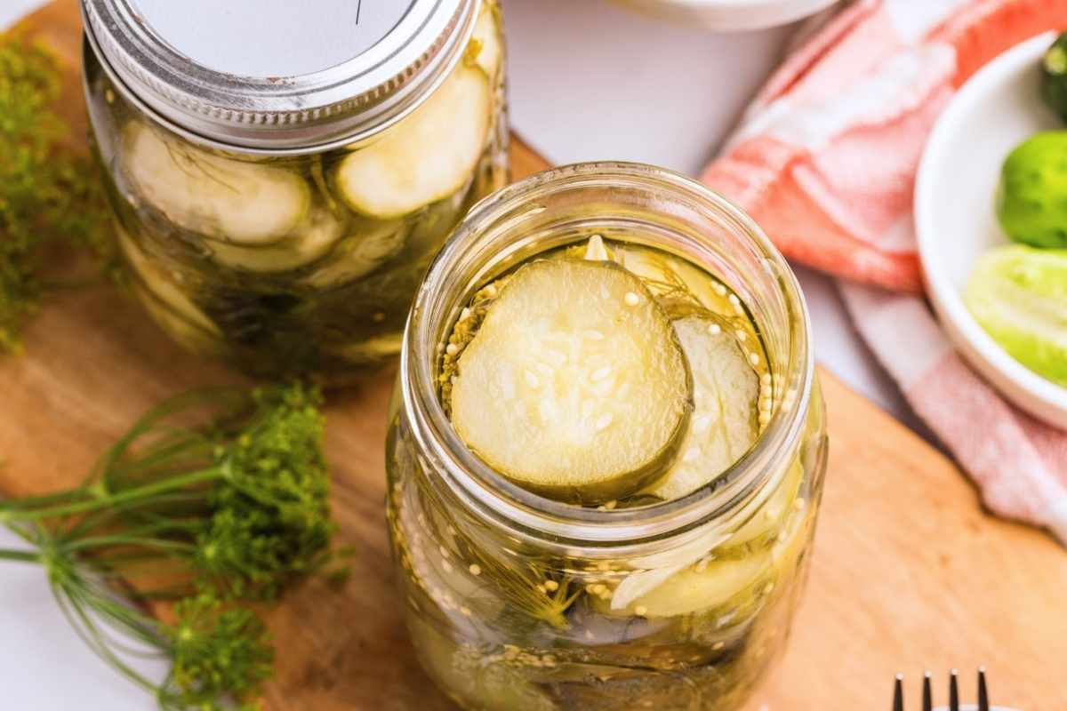 Two mason jars of sliced dill pickles on a wooden board with fresh dill and parsley.