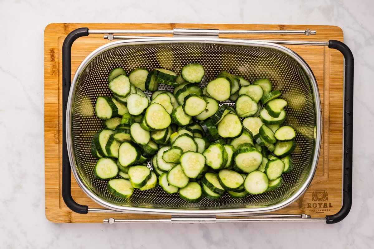Sliced cucumber rounds draining in a metal colander set on a bamboo cutting board.