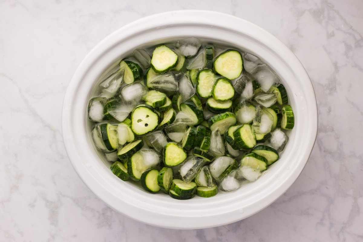 Sliced cucumbers soaking in ice water inside a white oval crock on a marble countertop.