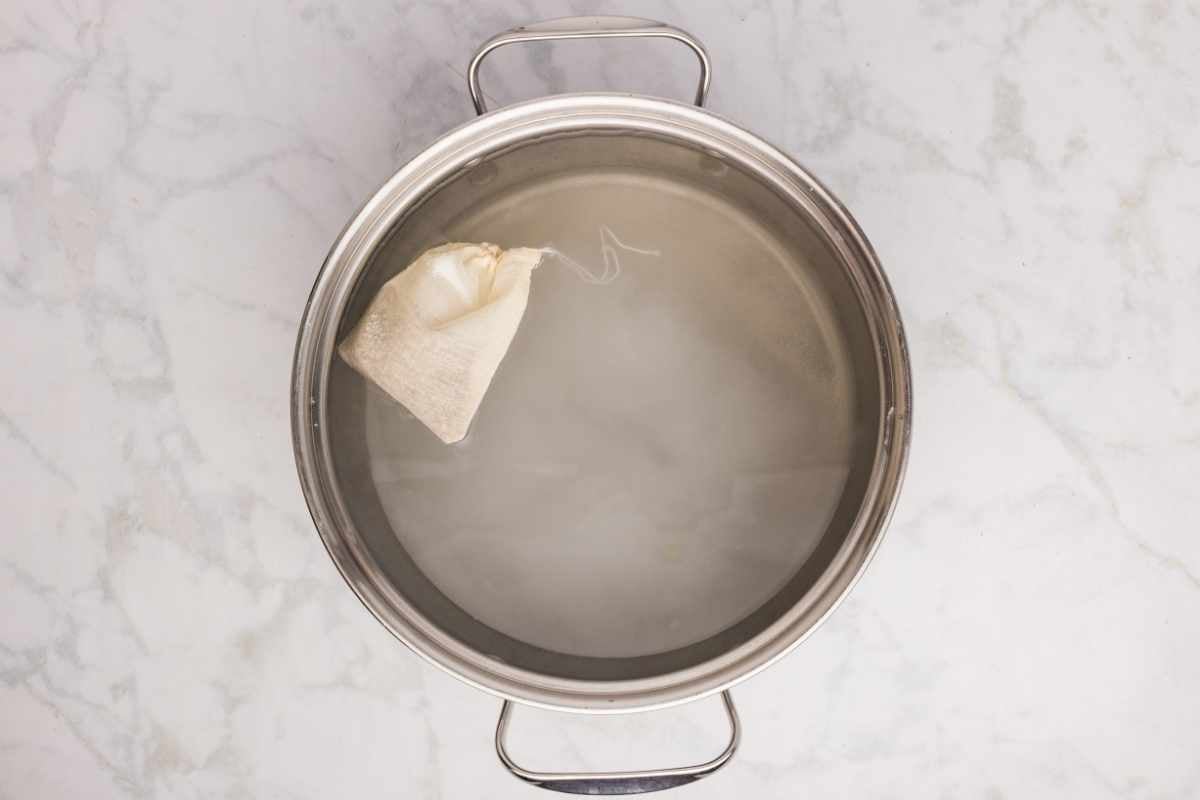 A muslin spice bag steeping in water inside a large stainless steel pot on marble.