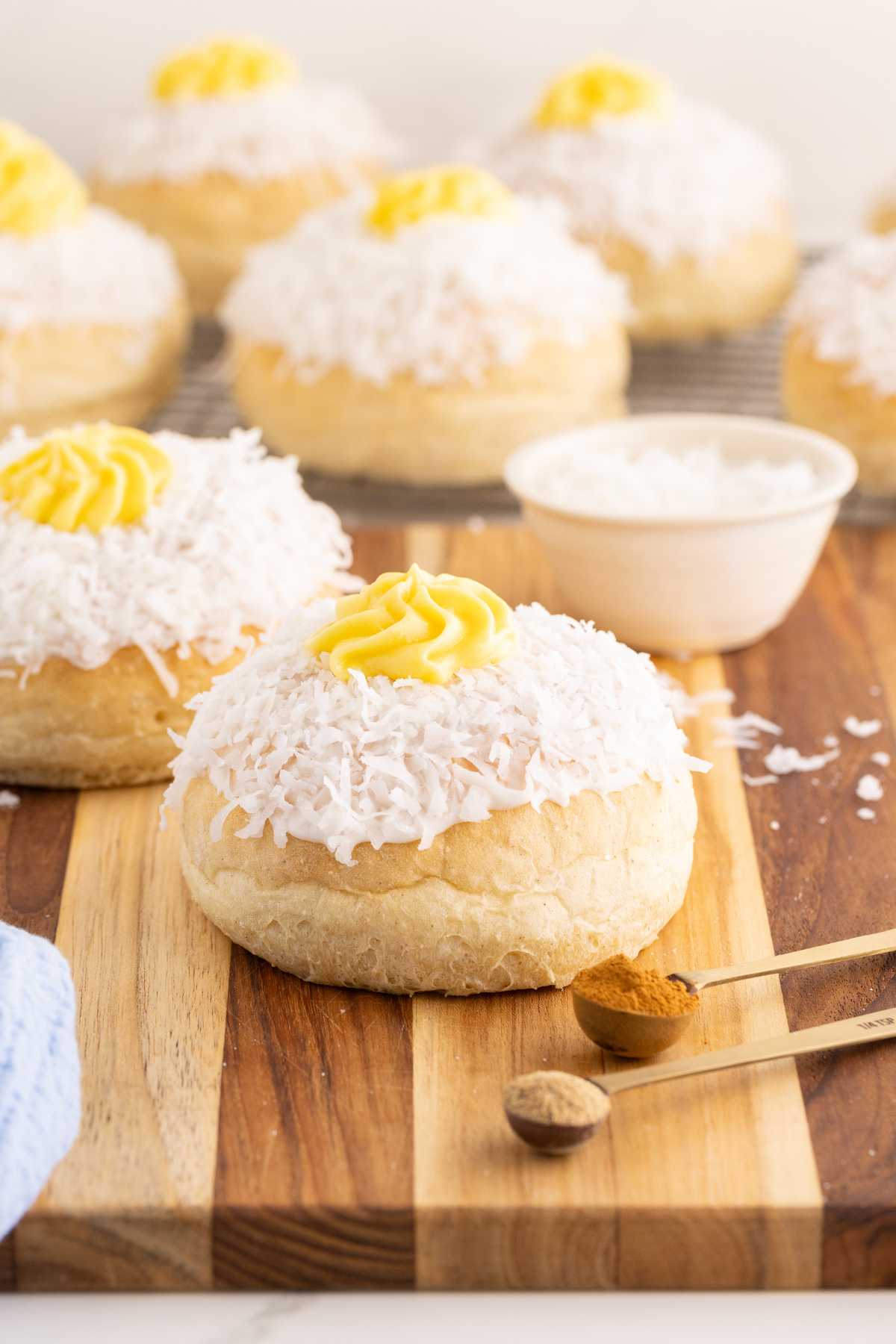 Coconut-coated school bread buns with yellow custard centers arranged on a wooden board with cinnamon-filled measuring spoons.