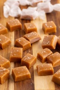 Square caramel candies topped with flaky sea salt scattered on a wooden cutting board with white wrappers behind.