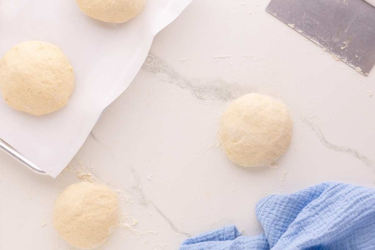 Round dough balls shaped on a floured marble counter with parchment, bench scraper, and blue towel nearby.