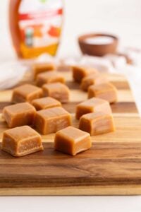 Squares of golden maple caramels arranged on a wooden cutting board with a syrup bottle behind.