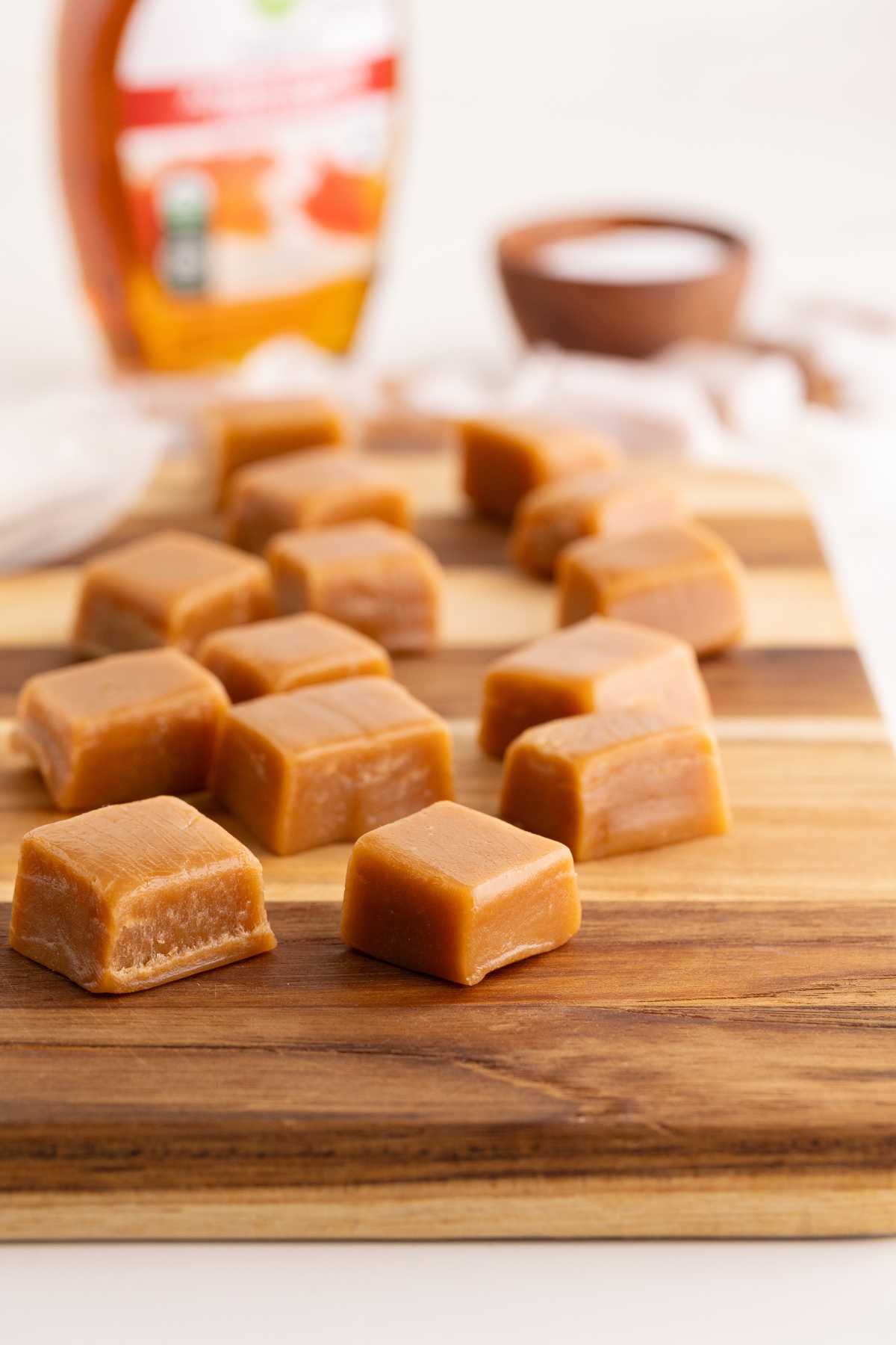 Squares of golden maple caramels arranged on a wooden cutting board with a syrup bottle behind.