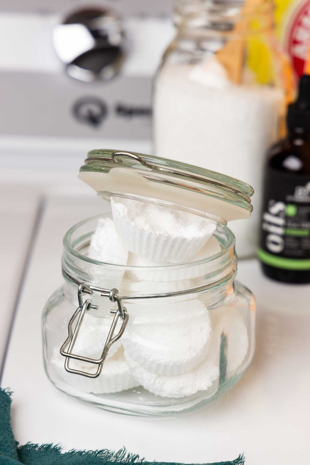 Homemade white washing machine tablets stacked in a clear glass jar beside essential oils and detergent.