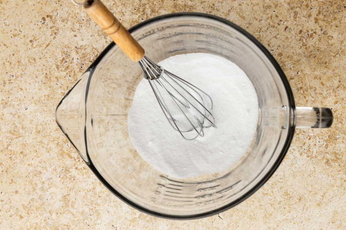 A wooden-handled whisk combining white powdered ingredients in a glass measuring bowl on a stone countertop.