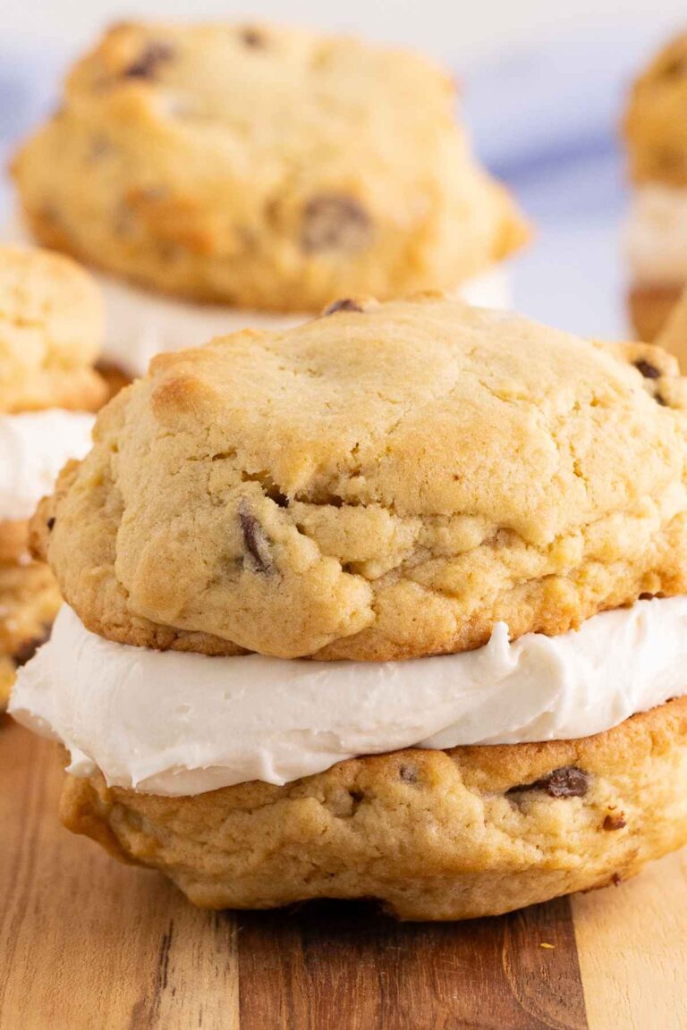 Chocolate chip cookie sandwiches filled with thick white cream frosting resting on a wooden board.