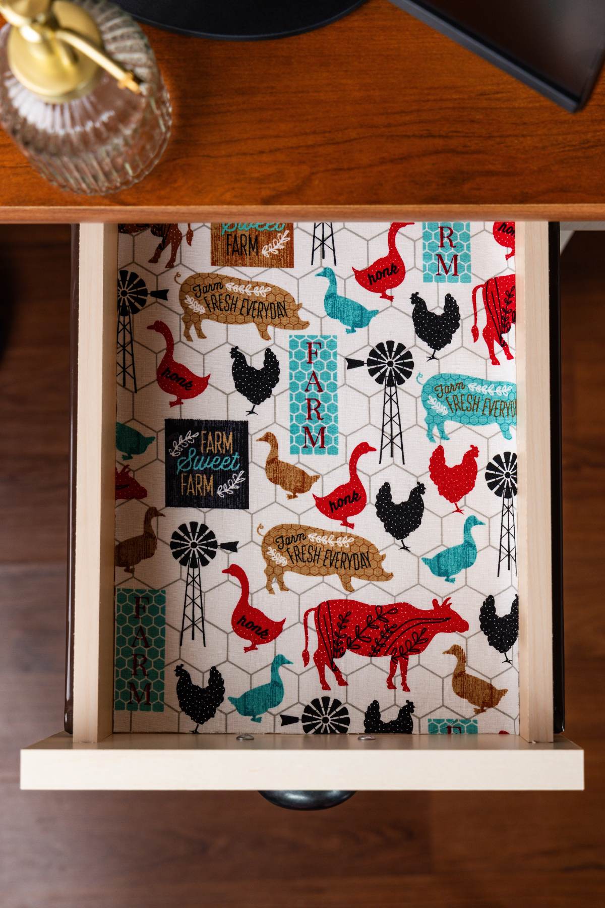 Open wooden drawer lined with farmhouse-print fabric featuring red cows, chickens, pigs, ducks, and windmills.