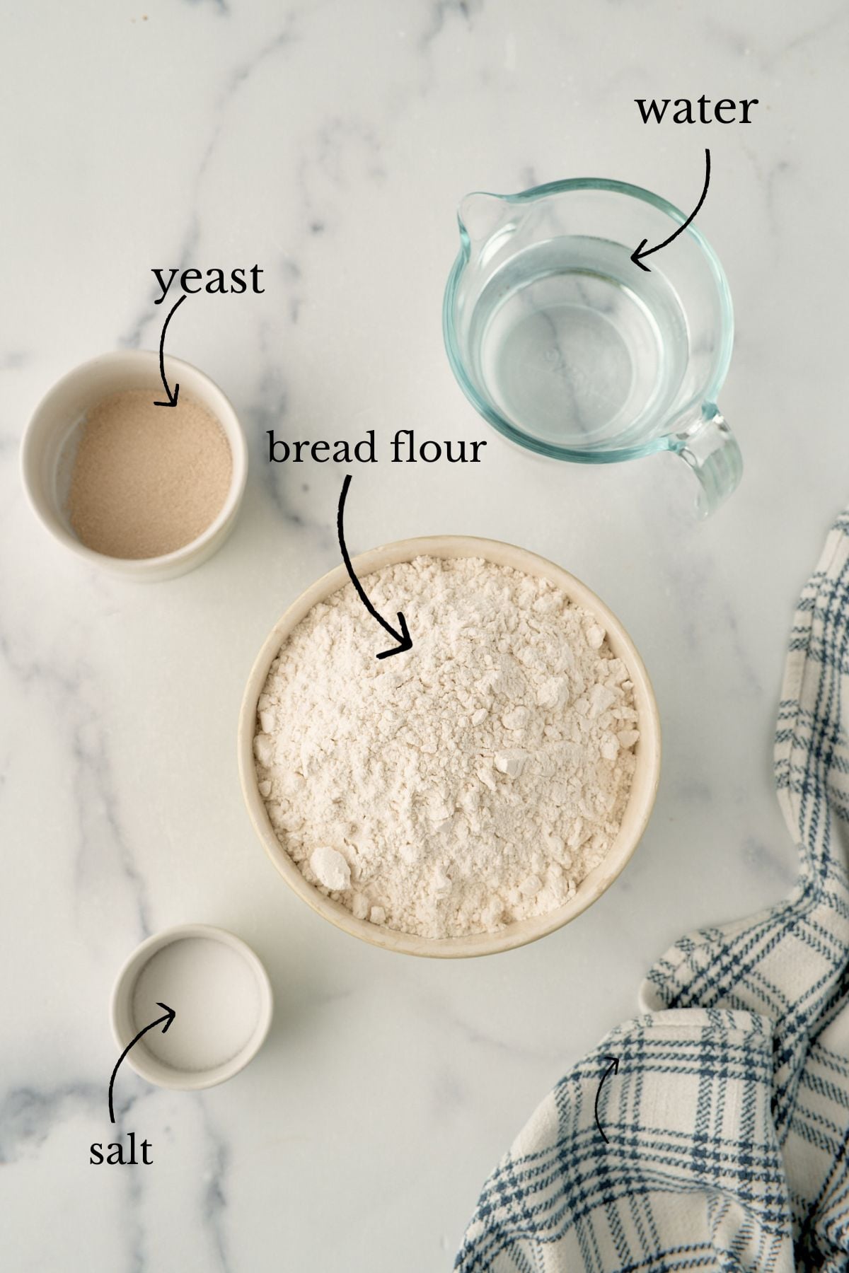 Bowls of bread flour, yeast, and salt beside a glass measuring cup of water on marble.