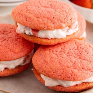 Pink strawberry whoopie pies stacked on a plate with creamy white filling and berry pieces peeking out.