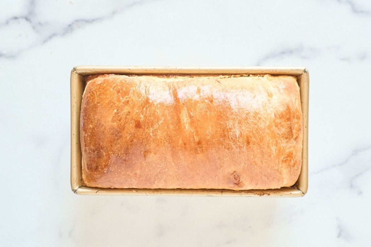 Freshly baked golden-brown sourdough loaf resting in a gold metal pan on white marble.