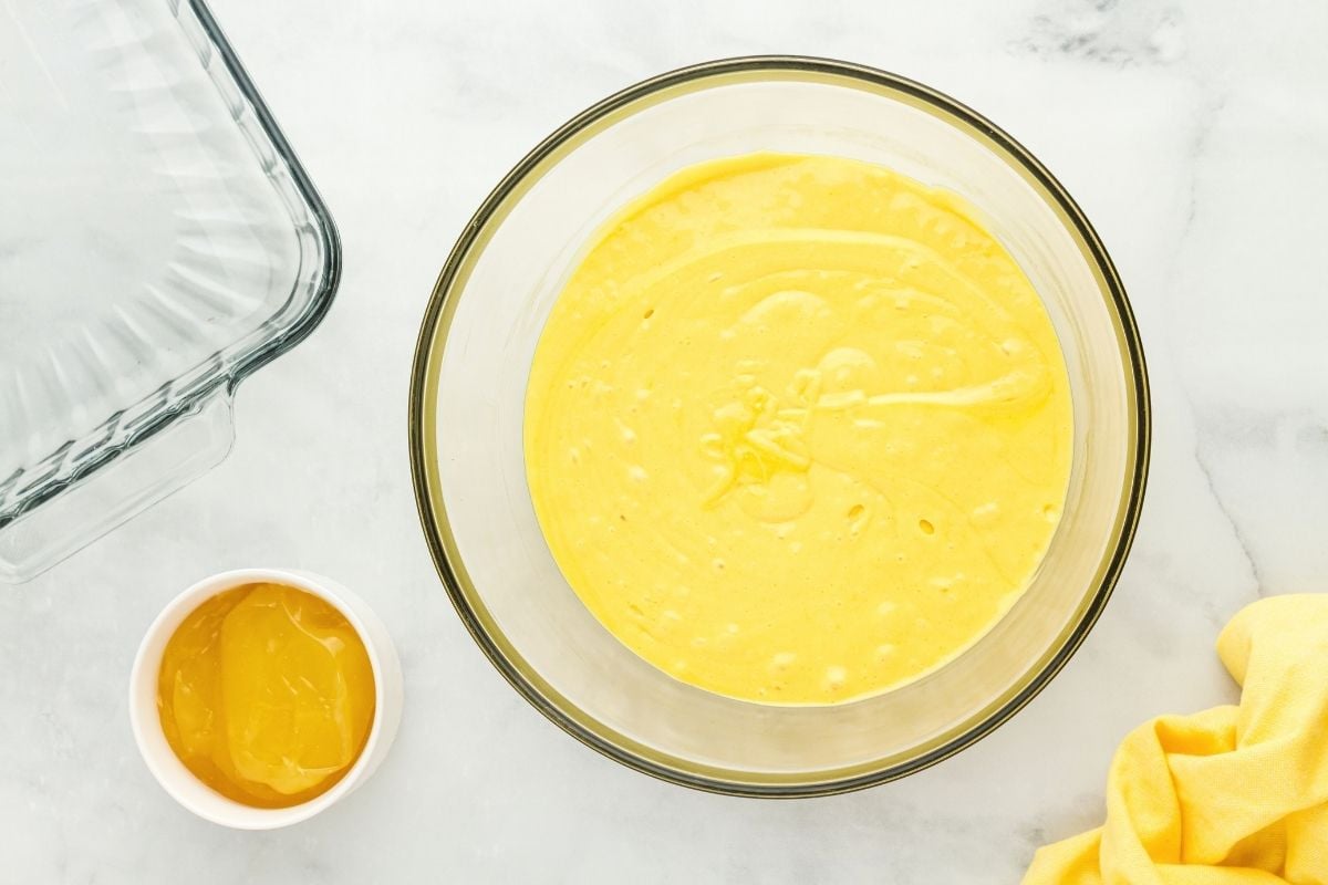 Smooth yellow lemon cake batter mixed in a glass bowl beside lemon curd and baking dish.