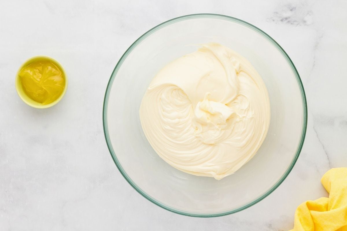 Whipped cream cheese frosting swirled smooth in a glass bowl beside a small dish of lemon curd.