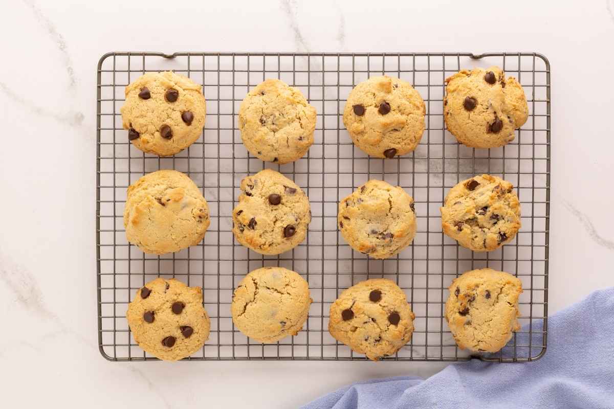 Twelve golden chocolate chip cookies cooling on a wire rack beside a blue linen napkin.