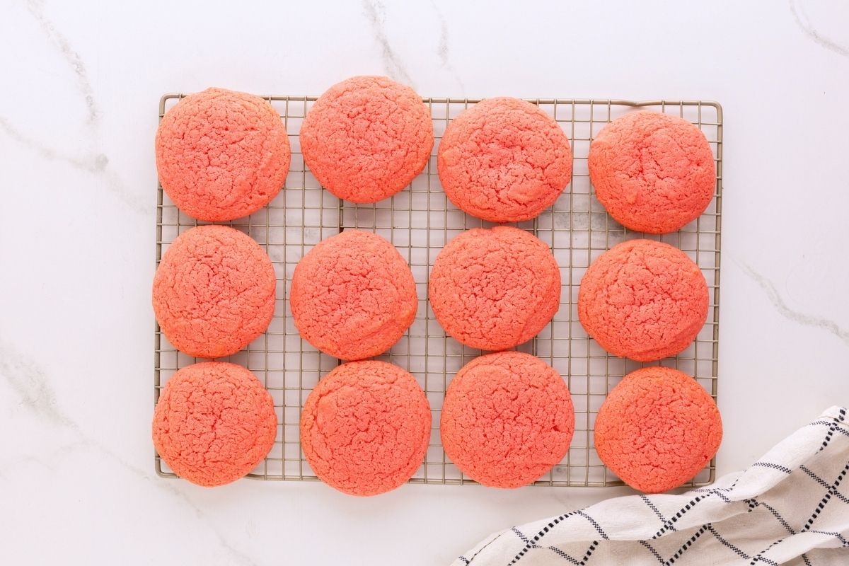 Twelve pink strawberry cookies cooling on a wire rack atop a white marble counter beside a striped towel.
