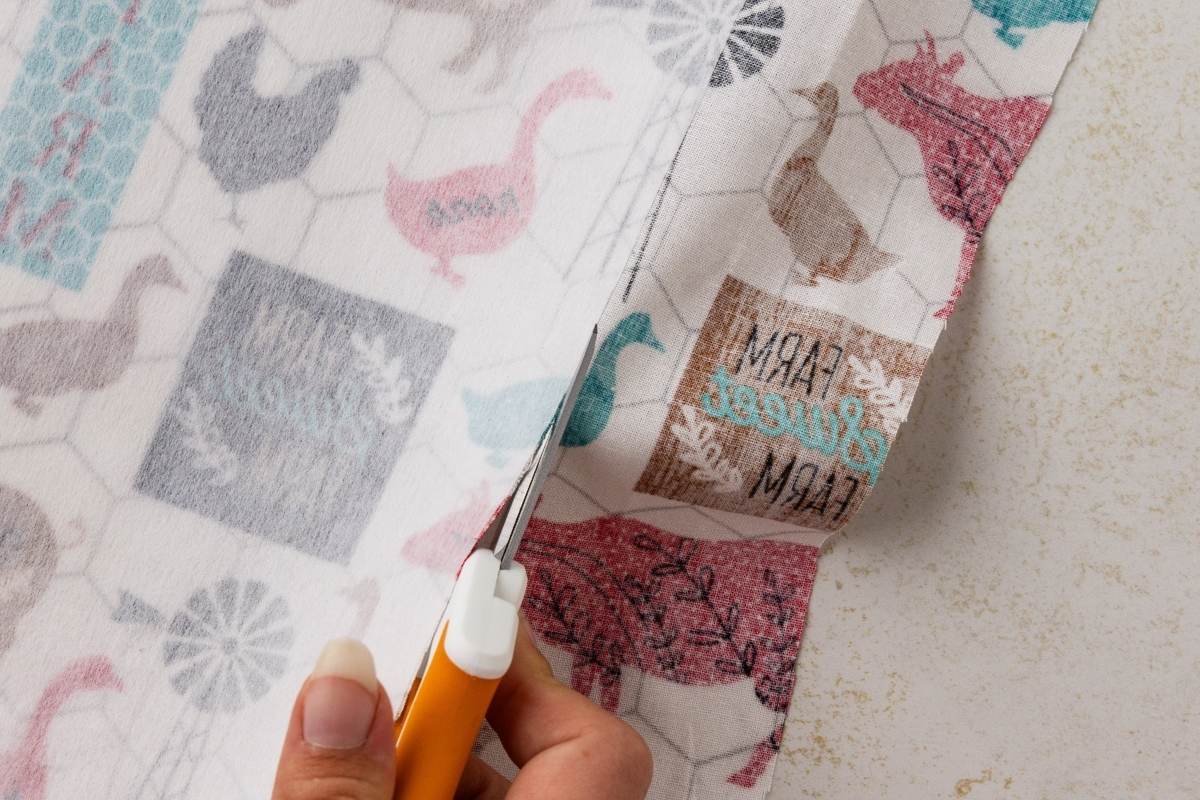 Hand using orange scissors to trim the edge of farmhouse-print fabric featuring pink and gray chickens and ducks.