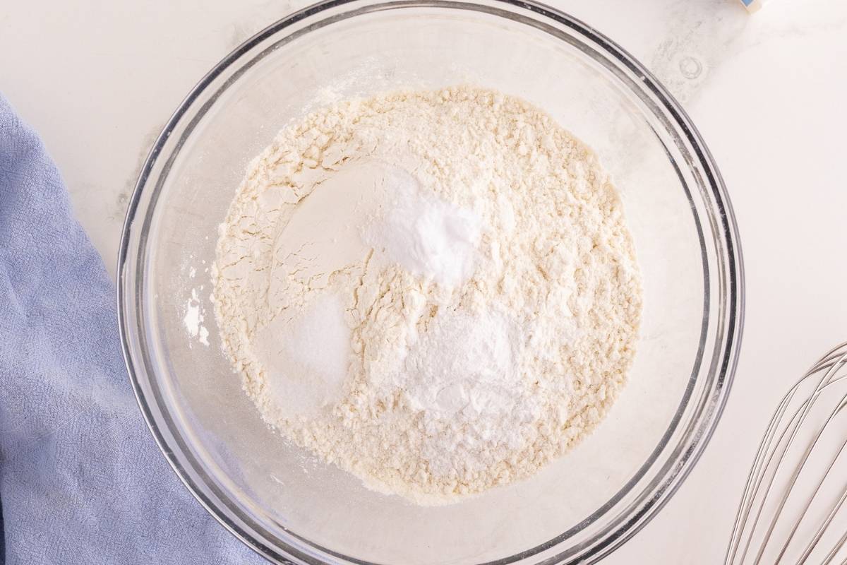 Flour, baking powder, baking soda, and salt combined in a glass mixing bowl beside a blue linen and whisk.