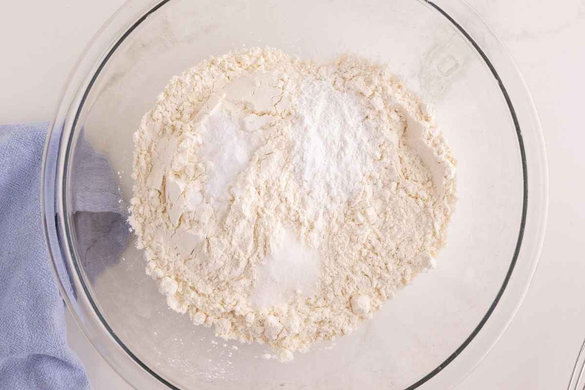 Flour, baking powder, and salt combined in a large glass mixing bowl beside a blue linen.