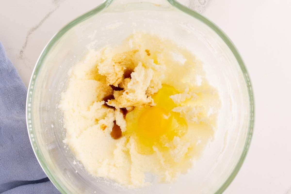 Cracked egg and vanilla extract added to creamed butter and sugar in a glass mixing bowl.
