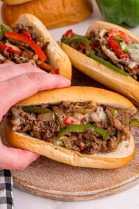 Hand holding a Philly cheesesteak loaded with steak, melted cheese, green peppers, and onions on a wooden board.