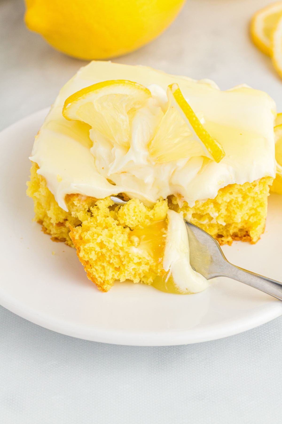 A slice of yellow lemon poke cake with creamy frosting and lemon slices, fork digging in.