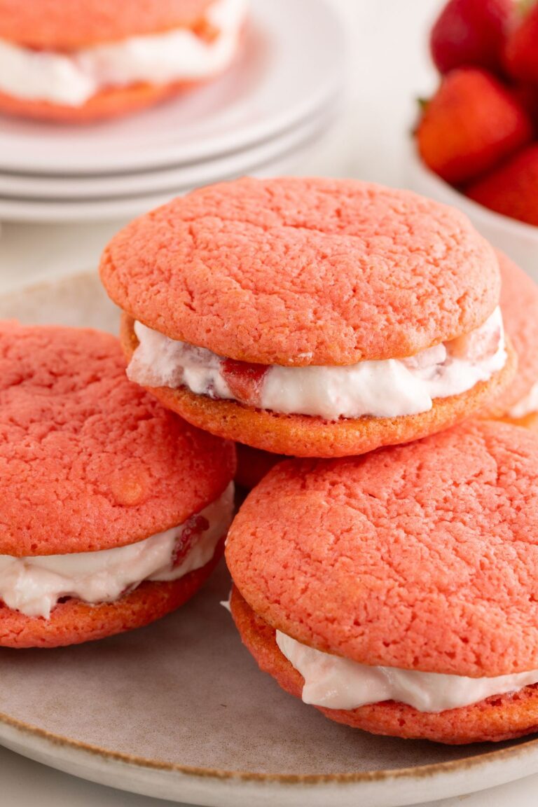 Pink strawberry whoopie pies stacked on a stoneware plate with cream filling and fresh strawberries nearby.