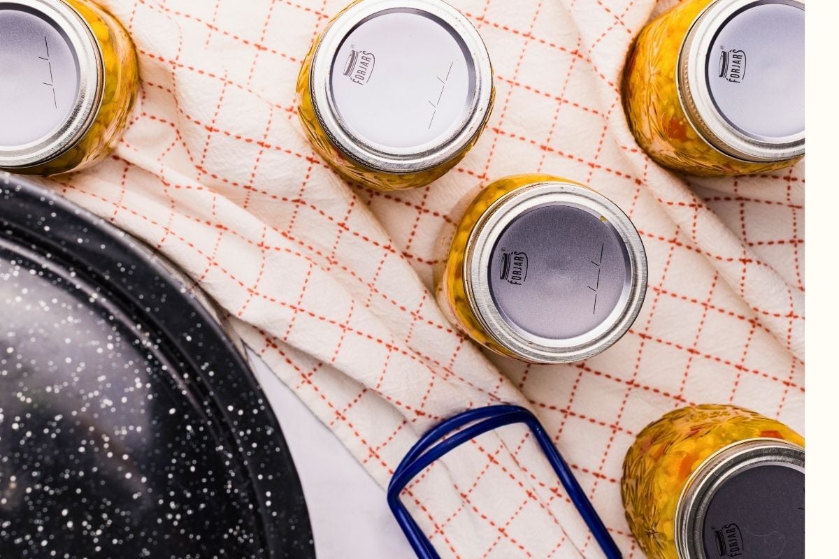 Overhead view of sealed jars of golden corn relish on a red-checked towel beside a speckled canning pot.