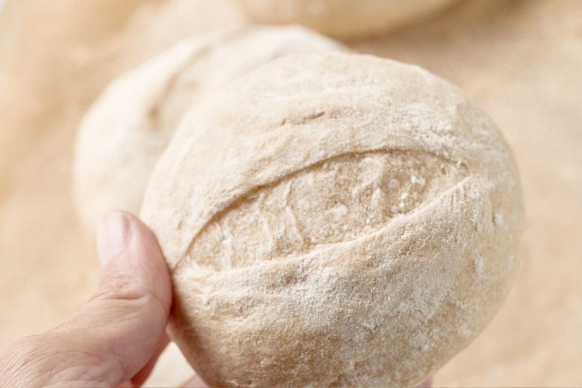 A hand holds a floured, scored round of French roll dough above a wooden surface.