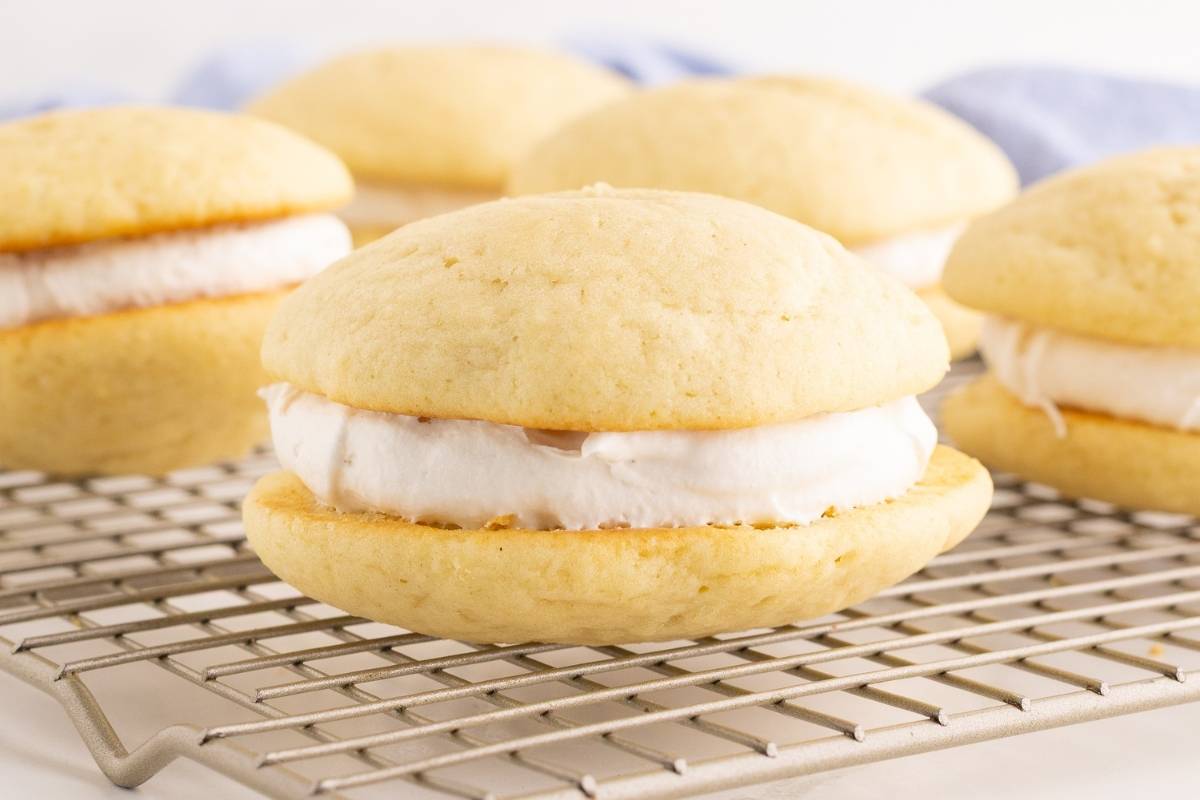Vanilla whoopie pies filled with fluffy white cream sit on a wire cooling rack near a blue linen napkin.
