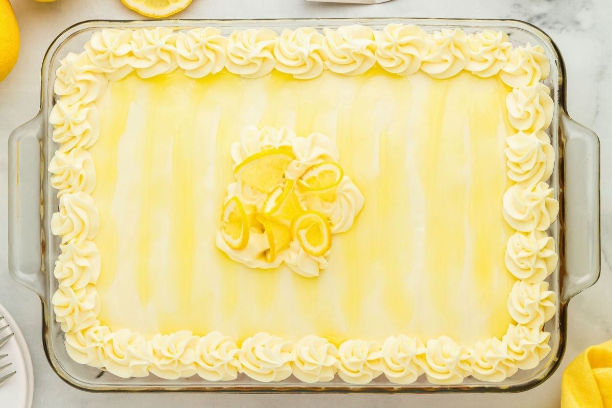 Glass baking dish filled with lemon poke cake frosted with piped buttercream rosettes and lemon slice garnish.