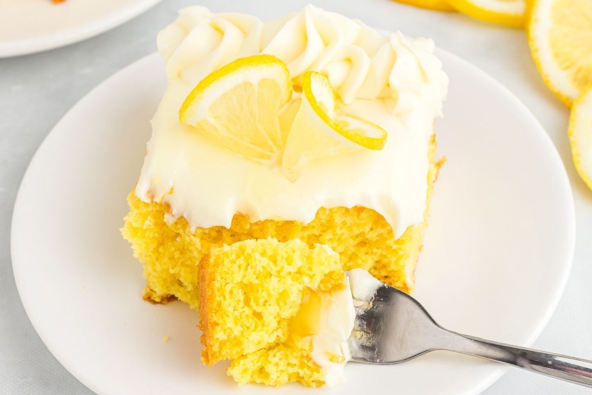 Yellow lemon poke cake slice topped with cream cheese frosting swirls and lemon twists on a white plate.