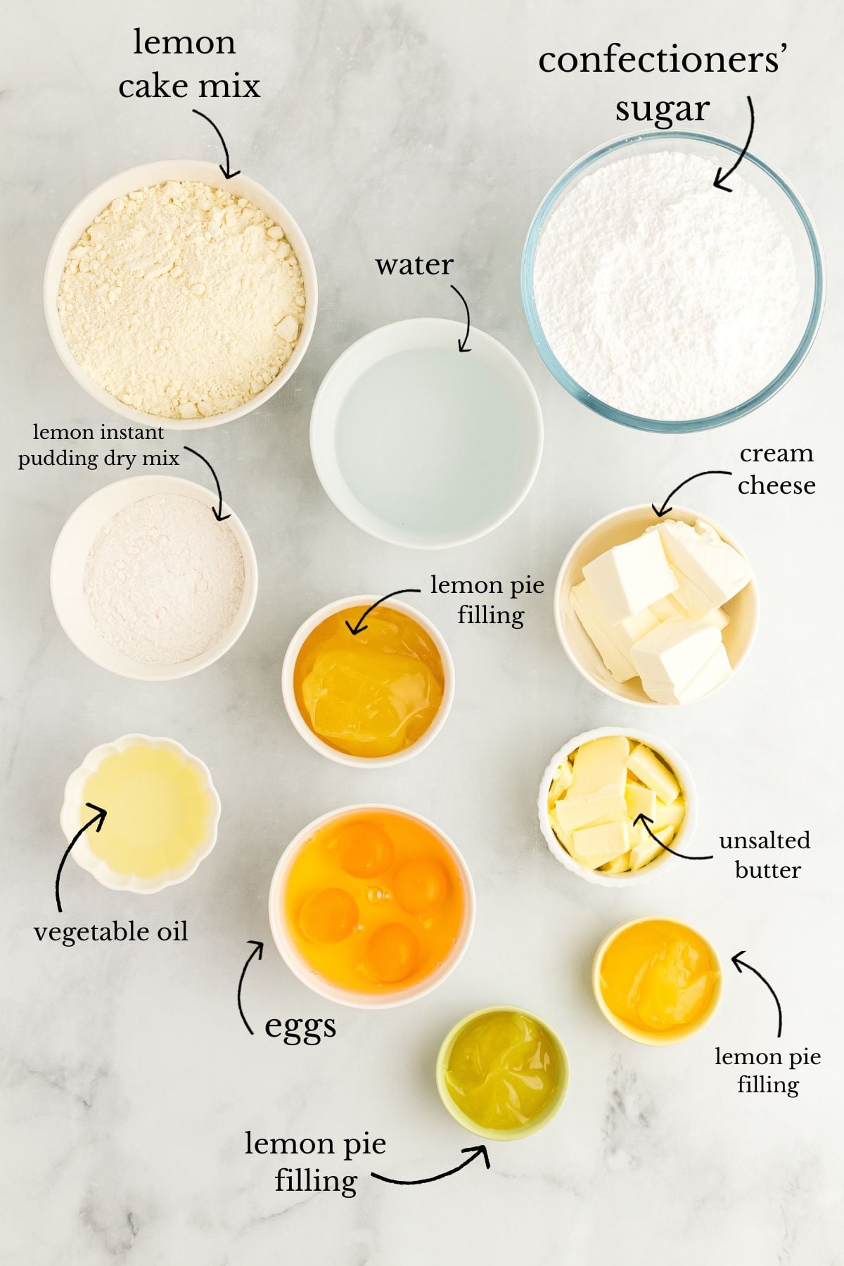Labeled bowls of lemon cake mix, pudding, eggs, lemon pie filling, cream cheese, butter, sugar, oil, and water on marble.
