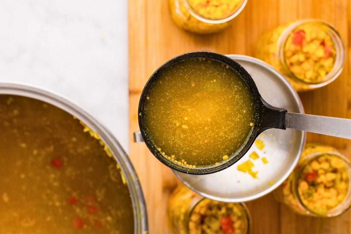 Ladle full of golden brine hovering over canning jars packed with corn relish on a wooden board.