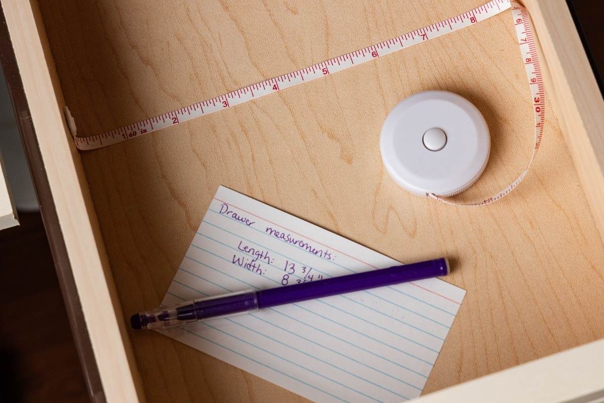 Measuring tape and index card noting drawer length and width inside an empty wooden drawer.