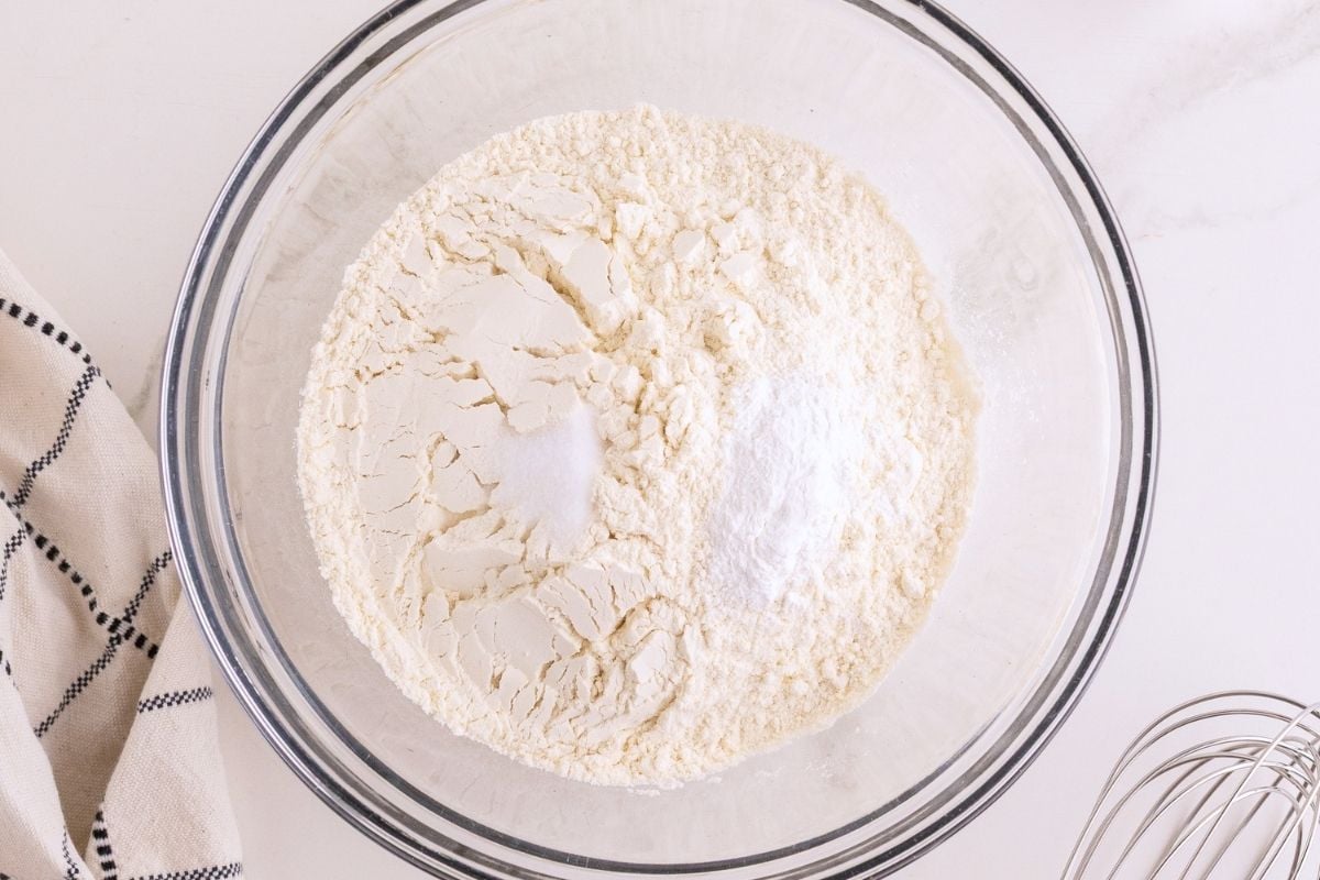 Flour, salt, and baking powder mounded in a glass mixing bowl beside a striped towel and whisk.
