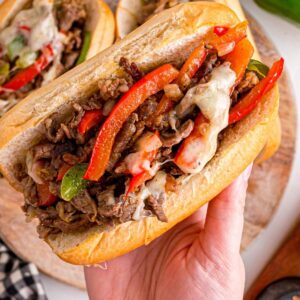 A hand holds a Philly cheesesteak sandwich loaded with steak, red and green peppers, onions, and melted cheese.