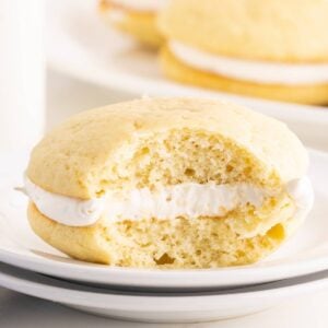 A vanilla whoopie pie with a bite taken out reveals fluffy white cream filling on a white plate.