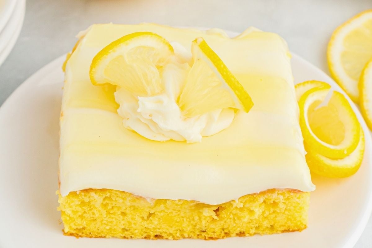 Square slice of yellow lemon poke cake topped with creamy frosting, whipped cream, and fresh lemon slices.