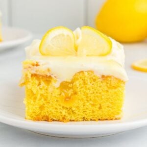 Slice of yellow lemon poke cake with cream cheese frosting topped with fresh lemon slices on white plate.