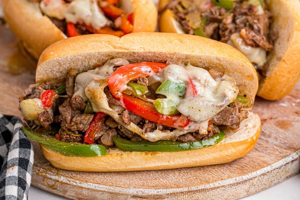 Philly cheesesteak on a hoagie roll filled with steak, melted cheese, red and green peppers on wooden board.