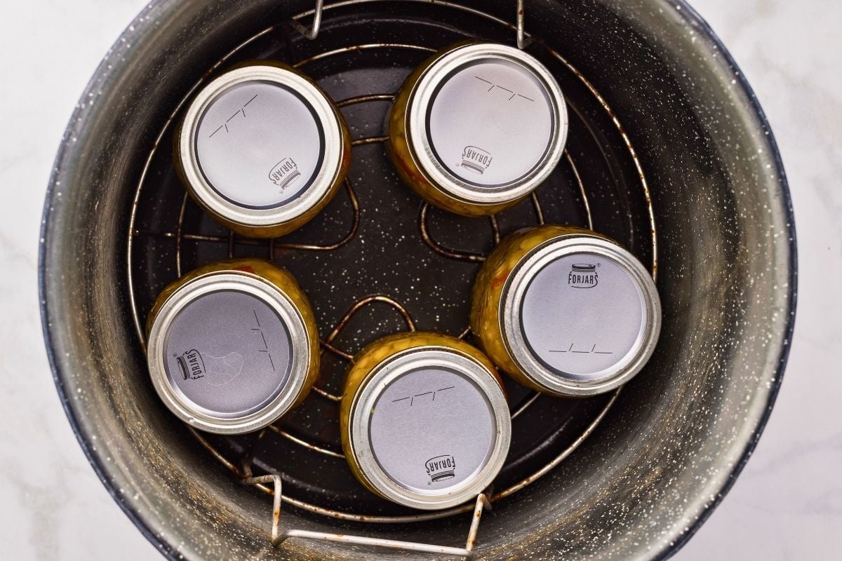Five sealed jars of corn relish arranged on a canning rack inside a speckled black pot.