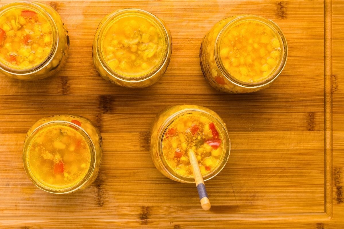 Wooden skewer removing air bubbles from jars of yellow corn relish on a bamboo cutting board.