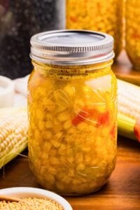 Sealed Ball mason jar of golden corn relish with red pepper pieces beside fresh corn cobs and mustard seeds.