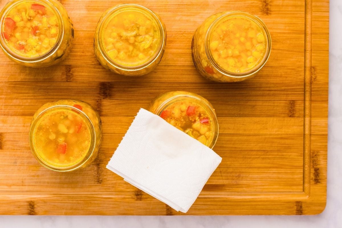 Five open jars of golden corn relish with red pepper pieces sitting on a wooden cutting board with a white cloth.